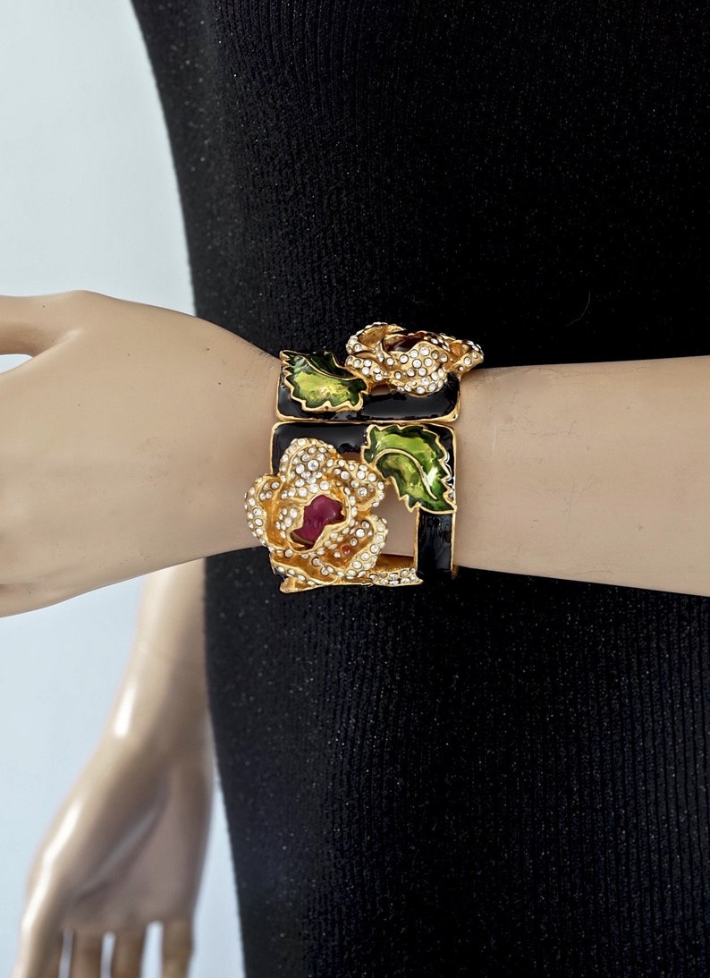 Puede incluir: Un brazalete de tono dorado con esmalte negro y pedrer&iacute;a. El brazalete presenta un dise&ntilde;o floral con hojas verdes y un centro rojo.