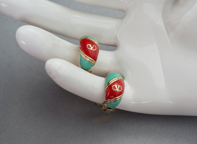 May include: A pair of gold hoop earrings with red and turquoise enamel. The earrings feature a gold logo in the center.