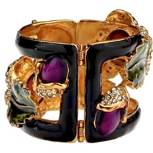 Puede incluir: Un brazalete de bisagra de esmalte negro con detalles dorados y detalles de esmalte de colores. El brazalete presenta un dise&ntilde;o floral con detalles de esmalte morado, verde y rojo. El brazalete est&aacute; adornado con pedrer&iacute;a transparente.