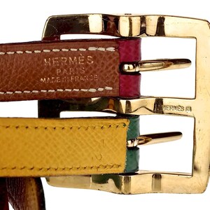 Pode incluir: Close-up de uma fivela de metal dourado com duas tiras de couro. A tira superior &eacute; marrom com costuras brancas e o texto "HERM&Egrave;S PARIS MADE IN FRANCE". A tira inferior &eacute; amarela com costuras verdes.
