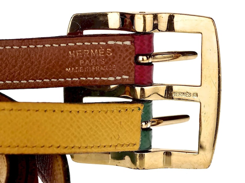 Pode incluir: Close-up de uma fivela de metal dourado com duas tiras de couro. A tira superior &eacute; marrom com costuras brancas e o texto "HERM&Egrave;S PARIS MADE IN FRANCE". A tira inferior &eacute; amarela com costuras verdes.