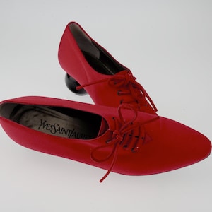 May include: A pair of red satin shoes with black laces and a round, black heel. The shoes have the text "YVES SAINT LAURENT" on the insole.