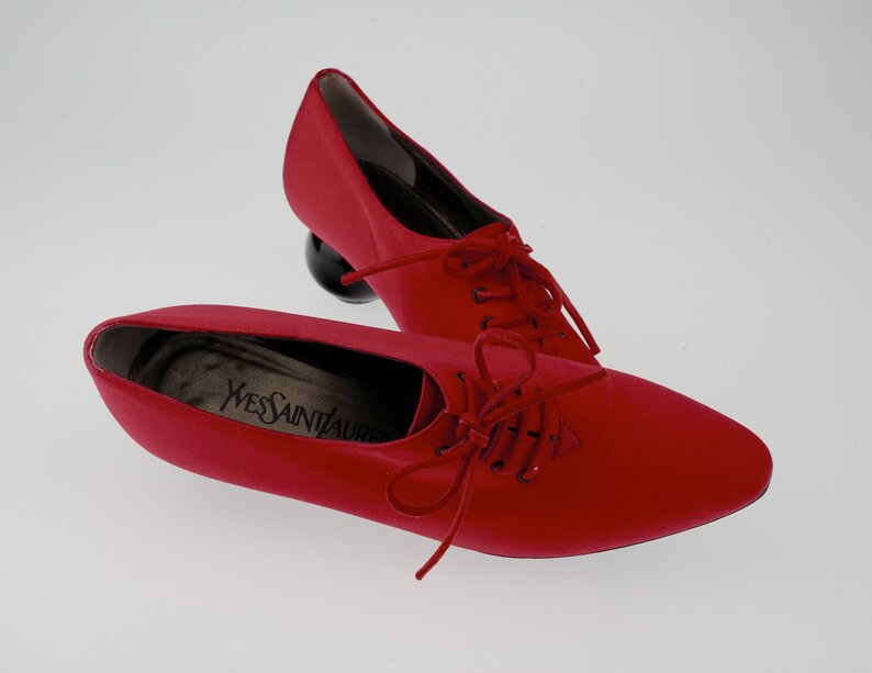 May include: A pair of red satin shoes with black laces and a round, black heel. The shoes have the text "YVES SAINT LAURENT" on the insole.