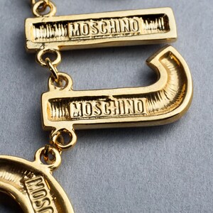 May include: Gold-tone necklace with multiple charms featuring the word "MOSCHINO" in a script font.