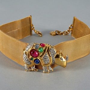 May include: A gold mesh choker necklace with a jeweled rhinoceros charm. The charm is adorned with rhinestones and colorful gemstones, including red, green, blue, and pink.