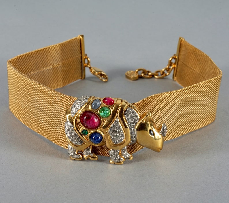 May include: A gold mesh choker necklace with a jeweled rhinoceros charm. The charm is adorned with rhinestones and colorful gemstones, including red, green, blue, and pink.