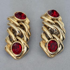 Pode incluir: Um par de brincos dourados com um design de elos de corrente. Cada brinco apresenta duas pedras preciosas ovais vermelho rubi. Os brincos têm uma estética vintage e um acabamento polido, adequados para ocasiões formais.