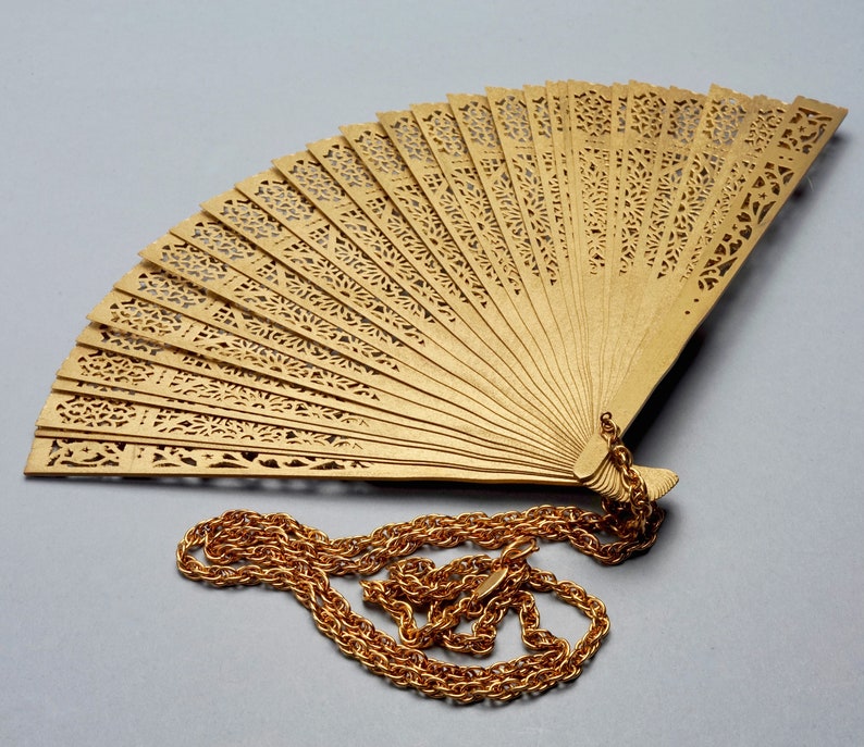 May include: A gold-colored, decorative fan with intricate cut-out designs. The fan is attached to a gold chain.