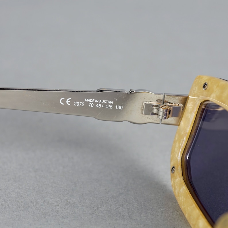 May include: Close-up of the silver metal arm of a pair of sunglasses with the text "CE 2972 70 46□25 130 MADE IN AUSTRIA" printed on the arm.