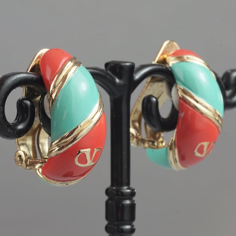 May include: A pair of red and turquoise clip-on earrings with gold accents. The earrings are shaped like a half-circle and have a logo on the front.