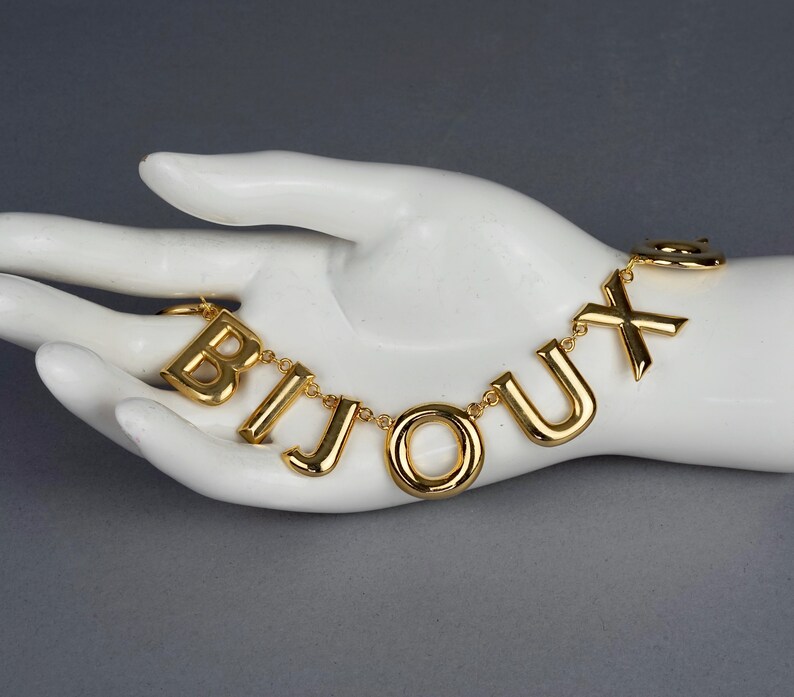 May include: A gold chain necklace with the word "BIJOUX" spelled out in large, chunky letters.