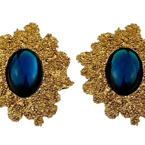 May include: A pair of gold tone earrings with a blue oval cabochon gemstone set in each. The earrings have a textured, floral design.