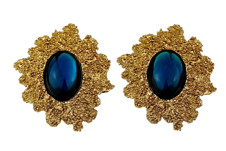 May include: A pair of gold tone earrings with a blue oval cabochon gemstone set in each. The earrings have a textured, floral design.