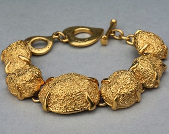 Puede incluir: Un brazalete dorado con un diseño texturizado y grueso. El brazalete está hecho de piezas ovaladas enlazadas con una superficie texturizada. El cierre es un cierre de palanca simple.
