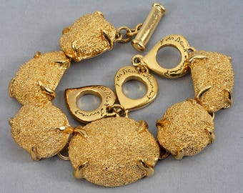 Puede incluir: Una pulsera dorada con eslabones redondeados y texturizados. La pulsera presenta un cierre de palanca y eslabones en forma de corazón con texto grabado. Los eslabones están conectados por pequeñas barras doradas. La pulsera está sobre un fondo gris.