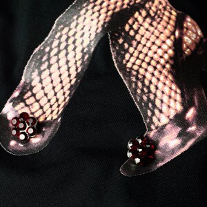 May include: Black t-shirt featuring a graphic of legs in fishnet stockings. The graphic includes red flower-shaped embellishments on the feet. The image has a dark, moody aesthetic.