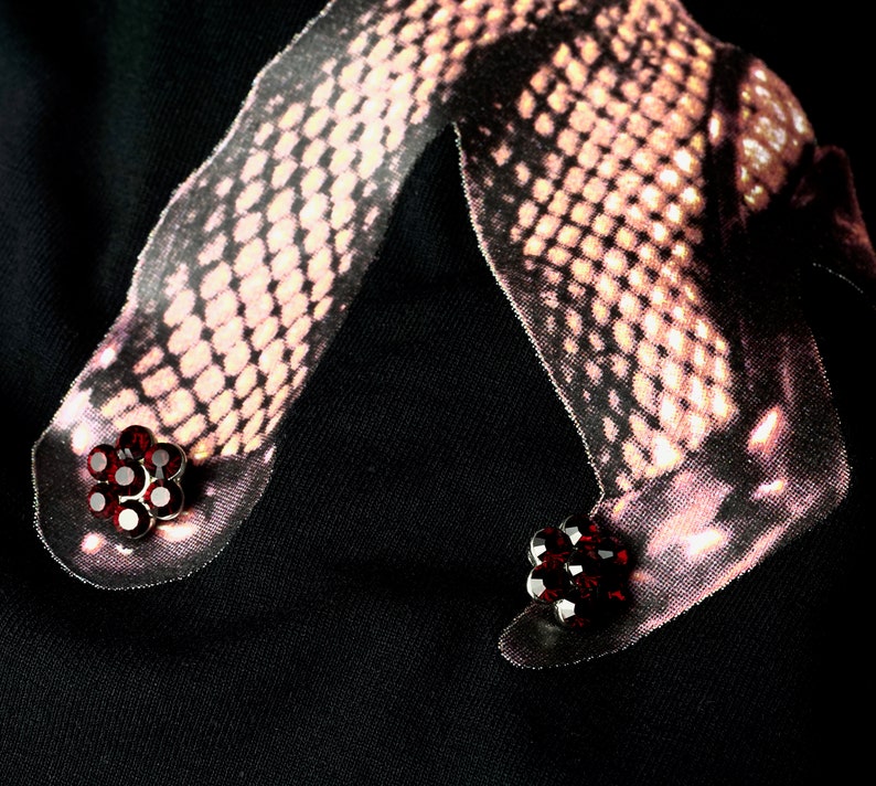 May include: Black t-shirt featuring a graphic of legs in fishnet stockings. The graphic includes red flower-shaped embellishments on the feet. The image has a dark, moody aesthetic.