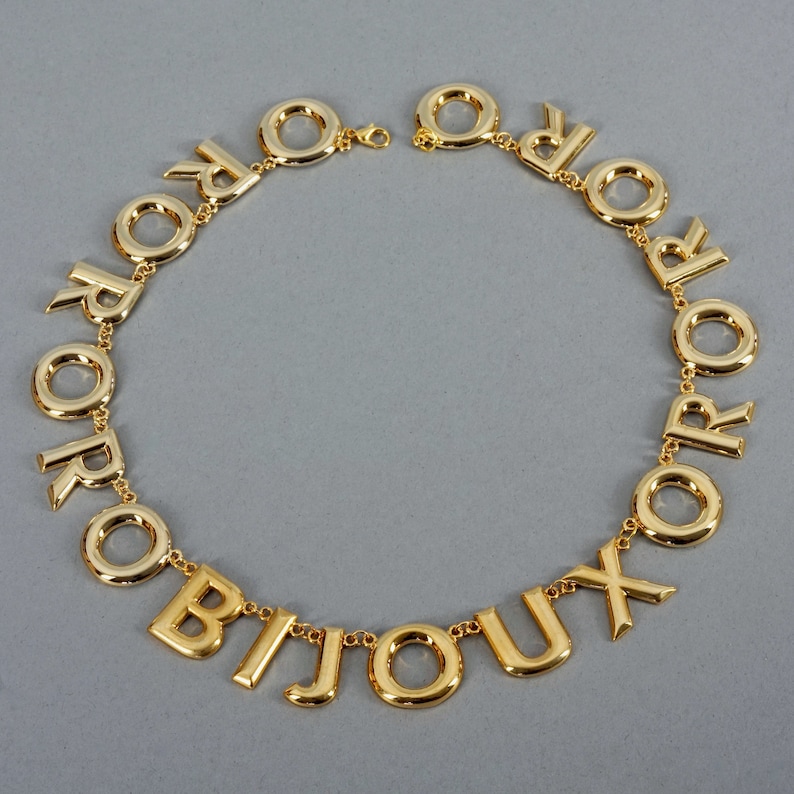 May include: A gold necklace with the word "BIJOUX" spelled out in large letters. The letters are connected by a chain with small gold circles.