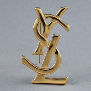 Puede incluir: Un broche de metal dorado con el icónico logotipo de YSL. El logotipo es un diseño estilizado de 'Y' y 'S' entrelazados.