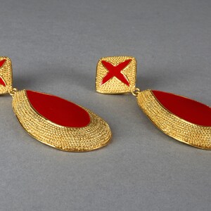 May include: A pair of gold tone dangle earrings with red enamel accents. The earrings feature a teardrop shape with a textured surface and a square top with a red cross design.