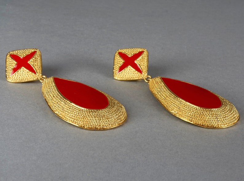 May include: A pair of gold tone dangle earrings with red enamel accents. The earrings feature a teardrop shape with a textured surface and a square top with a red cross design.