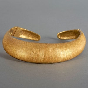 Könnte beinhalten: Ein goldfarbenes, offenes Armreif-Armband mit strukturierter Oberfläche. Das Armband hat ein gebogenes, abgerundetes Design und ist an den Enden offen. Das Armband besteht aus massivem Metall und hat ein gebürstetes Finish.