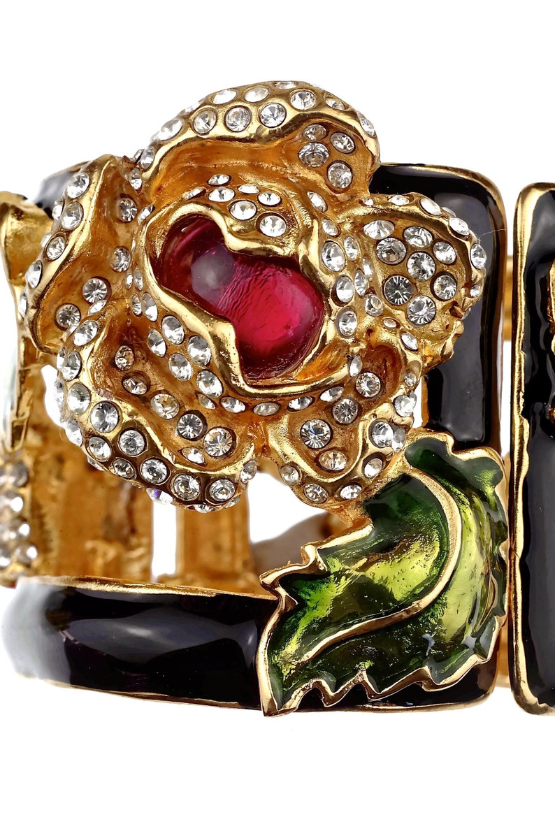 Puede incluir: Un brazalete de tono dorado con base de esmalte negro y un gran dise&ntilde;o de flor enjoyada. La flor est&aacute; engastada con diamantes de imitaci&oacute;n transparentes y tiene una piedra central roja.