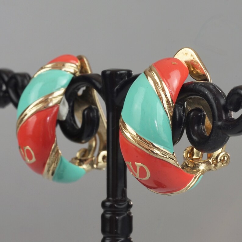 May include: A pair of gold tone clip-on earrings with a red and turquoise enamel design. The earrings are shaped like a half-circle and have a gold tone band around the edge.