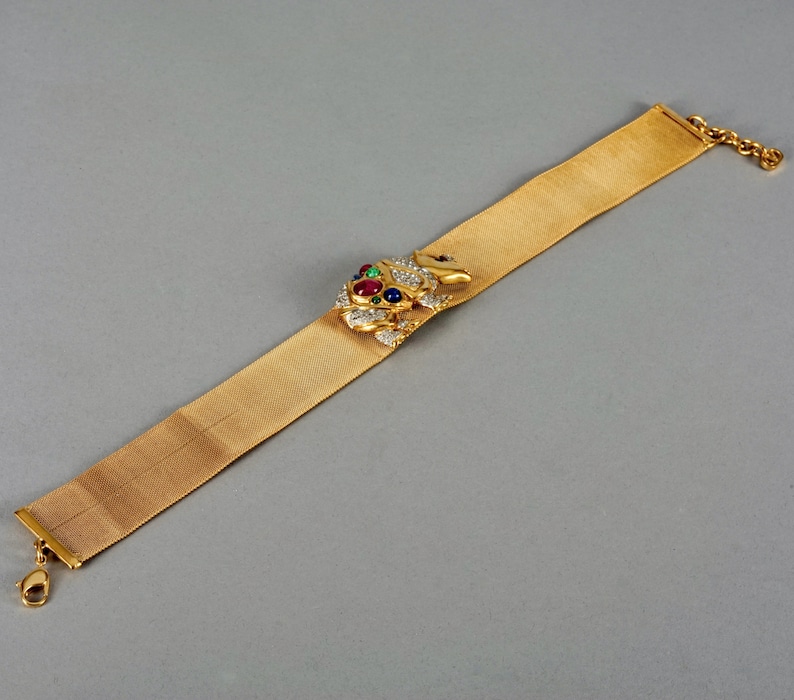May include: A gold mesh bracelet with a jeweled frog charm. The frog is adorned with red, green, and blue gemstones.