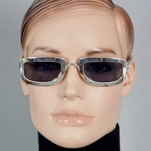 May include: Silver framed sunglasses with dark gray lenses. The sunglasses have a rectangular shape with rounded corners.