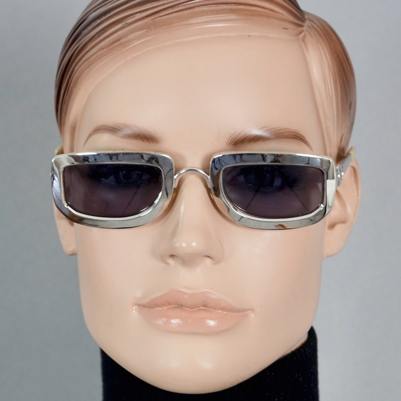 May include: Silver framed sunglasses with dark gray lenses. The sunglasses have a rectangular shape with rounded corners.