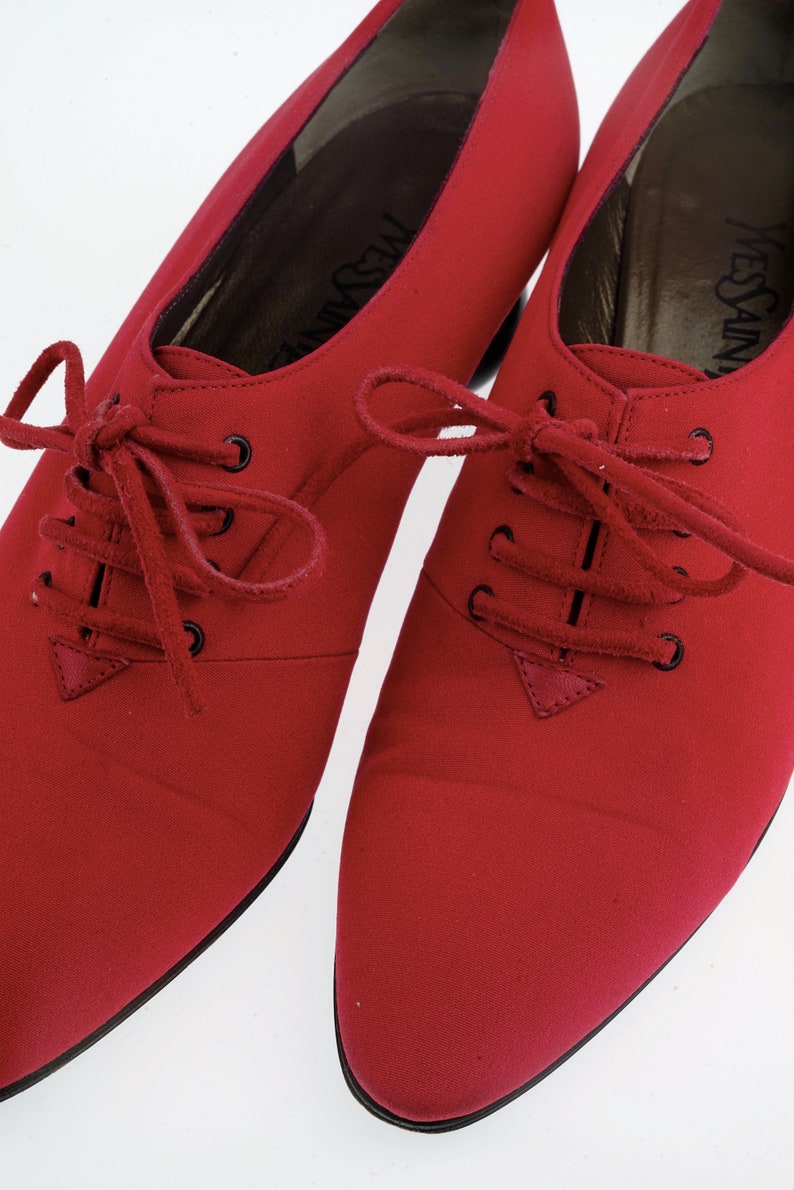 May include: A pair of red leather lace-up shoes with pointed toes. The shoes have a black leather sole and a black leather heel. The shoes are made by Yves Saint Laurent.