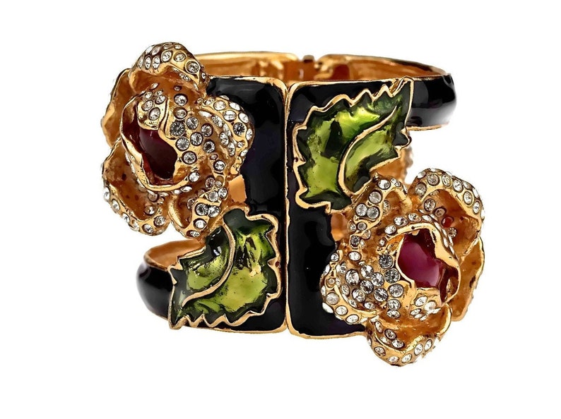 Puede incluir: Un brazalete r&iacute;gido de tono dorado con detalles florales en esmalte negro y verde. El brazalete est&aacute; decorado con pedrer&iacute;a transparente y piedras cabuj&oacute;n rojas y rosas.