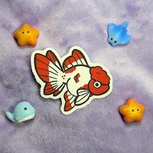 May include: A sticker of a red and white goldfish surrounded by small, colorful sea creatures. The sea creatures include two orange starfish, a blue stingray, and a light blue whale. The background is a soft, purple fabric.