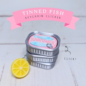 May include: A silver-colored tinned fish keychain clicker featuring a pink top with two light blue fish and the text SARDINES. A yellow lemon slice charm is attached to the side. The text TINNED FISH KEYCHAIN CLICKER and CLICK! appear.