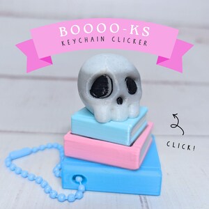 May include: A white glittery skull sits atop a stack of three books in light blue and pink. The object is a BOOOO-KS KEYCHAIN CLICKER with a light blue ball chain attached to the bottom book. A black arrow points to the side with the text CLICK!.