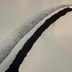 May include: Abstract art piece featuring a curved design with a black, white, and gray color scheme. The composition includes a black strip bordered by white and gray textured layers, set against a white background. The artwork has a minimalist aesthetic.