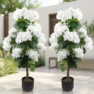 May include: Two artificial hydrangea trees with white flower clusters and green leaves. The trees have brown trunks and are planted in black, ribbed pots. The trees are placed on a light-colored patio.