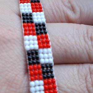 Könnte beinhalten: Ein Armband mit Perlen in einem Schachbrettmuster aus roten, schwarzen und weißen Perlen. Das Armband besteht aus kleinen, runden Perlen, die zu einem flexiblen Band aufgereiht sind. Das Armband wird am Handgelenk einer Person gezeigt.