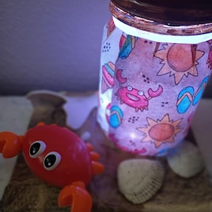 Könnte beinhalten: Ein von innen beleuchtetes Glasgefäß mit einem umlaufenden Muster aus Krabben, Sonnen und Flip-Flops. Das Glas hat einen braunen Metalldeckel mit einer Sonnengrafik. Ein kleines rotes Krabbenspielzeug aus Kunststoff und zwei weiße Muscheln liegen davor.