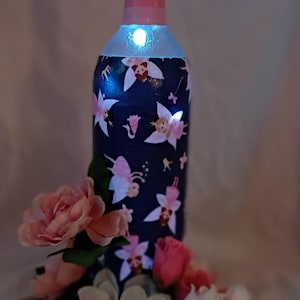 May include: A decorative wine bottle with a fairy design, illuminated with a soft glow. The bottle is adorned with pink and white flowers. The bottle's measurements are approximately 12 inches tall.