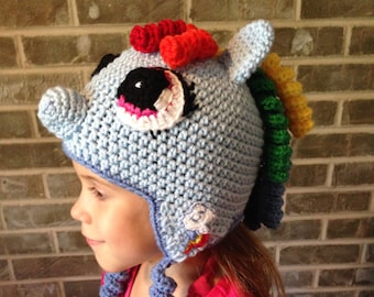 May include: A blue crocheted hat with a rainbow mane and a unicorn horn. The hat has a large eye with black and white details. The hat is worn by a child wearing a pink shirt.
