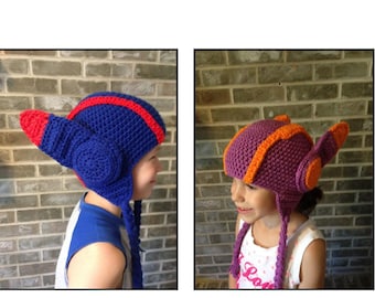 May include: Two children wearing crocheted hats shaped like helmets. The hat on the left is blue with red accents and the hat on the right is purple with orange accents.