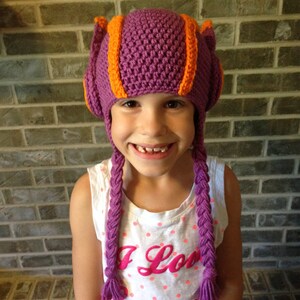 May include: A young person wearing a purple and orange crocheted hat with long braids. The hat has ear flaps and a pointed top. The person is wearing a white shirt with pink polka dots and the text "I Love Summer" printed on it.