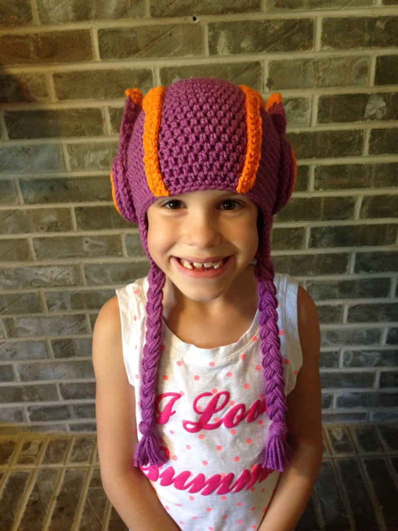 May include: A young person wearing a purple and orange crocheted hat with long braids. The hat has ear flaps and a pointed top. The person is wearing a white shirt with pink polka dots and the text "I Love Summer" printed on it.