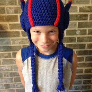 May include: A blue crocheted hat with red stripes and long blue braids. The hat is worn by a child with a grey and blue shirt.