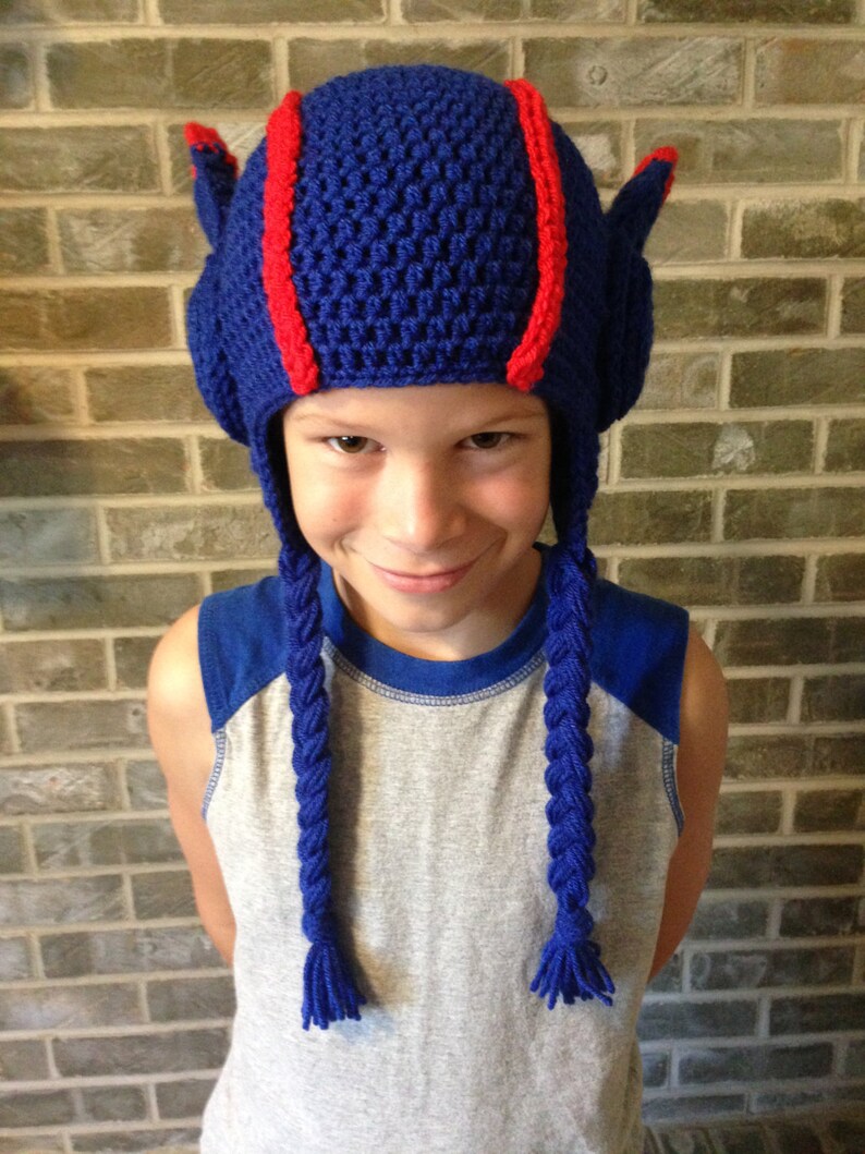 May include: A blue crocheted hat with red stripes and long blue braids. The hat is worn by a child with a grey and blue shirt.