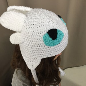 Puede incluir: Sombrero blanco de ganchillo con detalles de ojos azules y negros. El sombrero tiene dos largas orejeras blancas.
