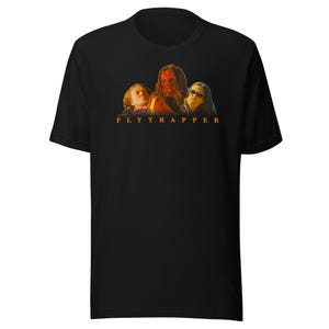 May include: Black t-shirt featuring a movie poster-style graphic with three figures and the text "FLYTRAPPER" in orange. The graphic has a warm color palette with orange and red tones.