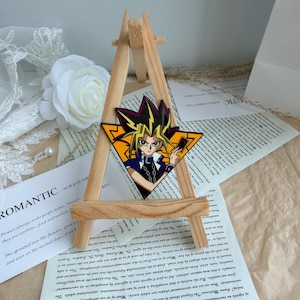 Könnte beinhalten: Eine dreieckige Emaille-Anstecknadel mit einer stilisierten Illustration der Anime-Figur Yugi Muto mit stacheligem gelb-schwarzem Haar, bekleidet mit einem blauen Hemd und einer silbernen Kette, vor einem leuchtend orangefarbenen Hintergrund mit schwarzen geometrischen Rändern.